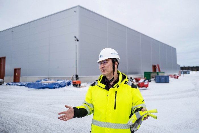Yun Lee , purtând o haină de înaltă vizibilitate și o cască de protecție, gesticulează cu mâinile în fața fabricii Northvolt