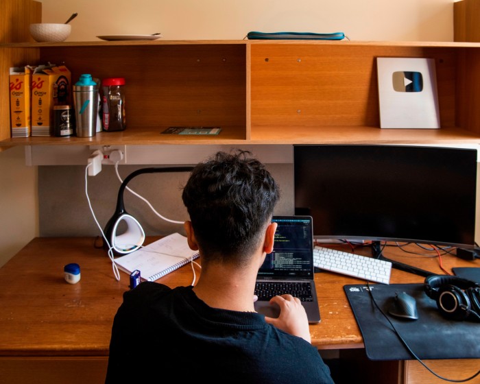Un tânăr, văzut din spate, stă la un birou și lucrează la un laptop